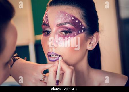 Après avoir peint le visage du modèle avec du violet et des cristaux, l'artiste de maquillage façonne ses lèvres avec un stylo de contour violet Banque D'Images