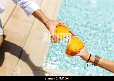 Vue de dessus les mains de couple dans une tenue décontractée assis près de la piscine ensemble et de boire des cocktails ou jus d'orange. Détendez-vous dans la piscine. Concept de vocation. Banque D'Images