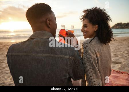 Un jeune couple multiethnique souriant boit du jus sur une plage de sable au coucher du soleil Banque D'Images