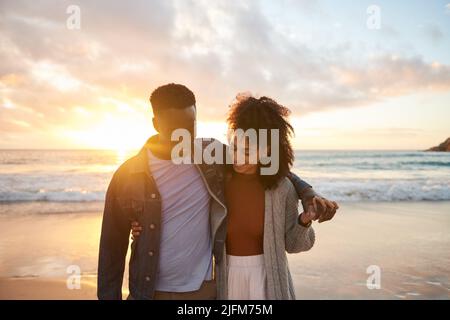 Couple multiethnique souriant marchant bras dans bras sur une plage au coucher du soleil Banque D'Images