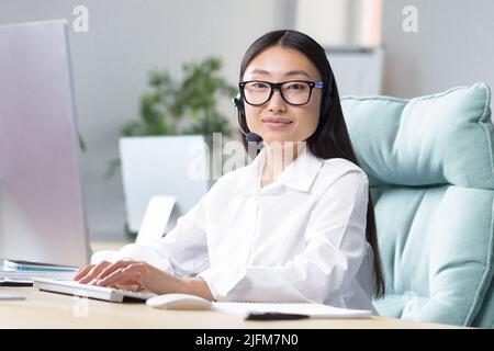 Jeune assistante technique, portrait d'une femme d'affaires asiatique travaillant dans un bureau moderne au travail, regardant la caméra, utilisant un casque pour la communication vidéo. Banque D'Images