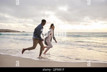 Jeune couple multiethnique qui court dans les vagues sur une plage Banque D'Images