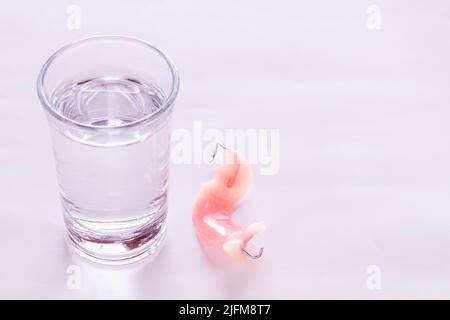 Gros plan des prothèses acryliques immergées et un verre d'eau, table dans la chambre. Arrière-plan simple et lumineux. Banque D'Images