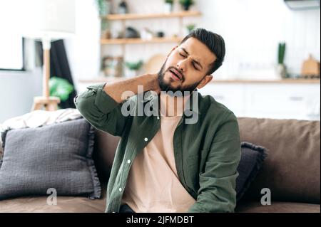 Un jeune homme indien ou arabe contrarié, assis sur un canapé dans un salon confortable, massant son cou, fatigué après une dure journée de travail, souffrant de problèmes articulaires, pincé nerf dans le cou, besoin de repos Banque D'Images