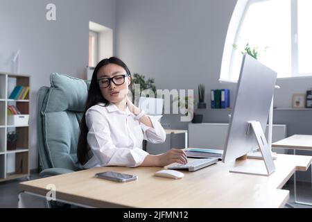 Femme asiatique surfatiguée employée de bureau, a de graves douleurs au cou, moque les muscles du cou avec la main, femme employée dans le bureau à l'ordinateur. Banque D'Images