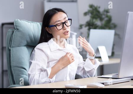 Chaleur dans le bureau, femme asiatique essayant de se rafraîchir, femme d'affaires travaillant à l'ordinateur, en agitant ses mains pour se refroidir. Banque D'Images