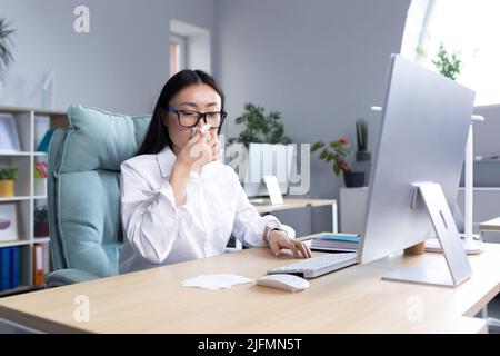 Une employée de bureau asiatique malade, a le froid et la grippe, tousse et éternue dans les tissus, travaille à l'ordinateur. Banque D'Images