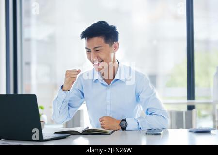 Un jeune homme d'affaires asiatique heureux et enthousiaste applaudit avec ses poings travaillant sur un ordinateur portable assis dans un bureau seul au travail. Un homme d'affaires chinois Banque D'Images