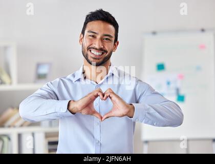 Jeune homme d'affaires de course mixte heureux faisant un geste de coeur avec ses mains debout dans un bureau seul au travail. Un patron hispanique masculin souriant montrant Banque D'Images