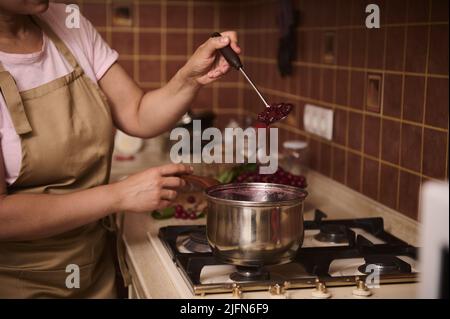 Gros plan d'une femme, d'une femme au foyer dans un tablier, debout sur la cuisinière et préparant de la confiture de cerises sur une casserole en métal dans la cuisine. Le conc Banque D'Images