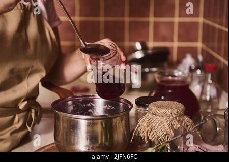 Gros plan d'une femme au foyer versant de la confiture de cerises maison dans des pots tout en se tenant à la cuisinière de la cuisine maison. Le concept de cuisine hom Banque D'Images