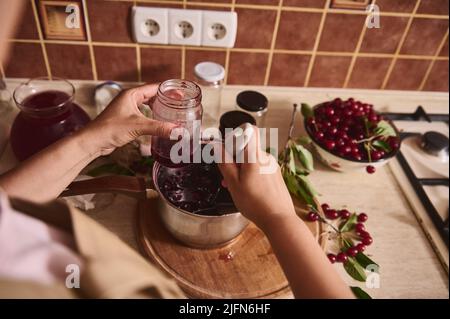 Vue rognée d'une femme au foyer, confiseur de femme debout près du comptoir de cuisine et verser de délicieux confits de cerise fraîchement brassés dans le steril Banque D'Images