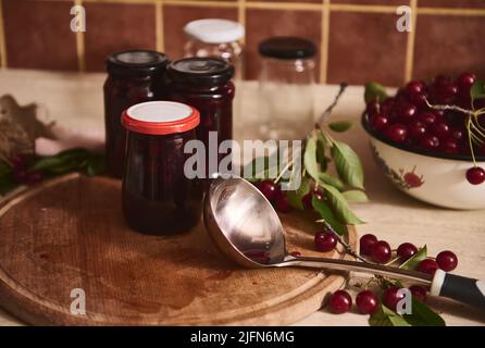 Confiture fraîchement préparée versée dans des pots en verre stérilisés se tient à côté d'une louche, baies de cerises vertes fraîches avec des feuilles vertes sur un panneau de bois dans un c rustique Banque D'Images