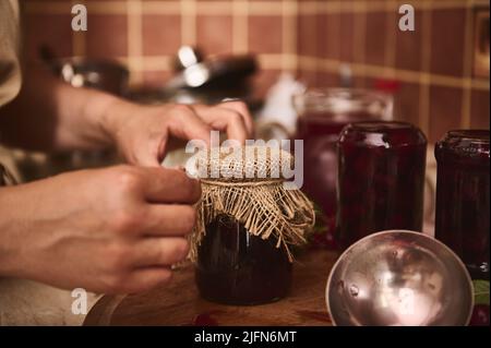 Concentrez-vous sur un pot de fruits frais confits et les mains de la femme de ménage mettant le couvre-bagages sur le couvercle du pot et nouant l'arc avec une corde tout en préparant la collecte Banque D'Images
