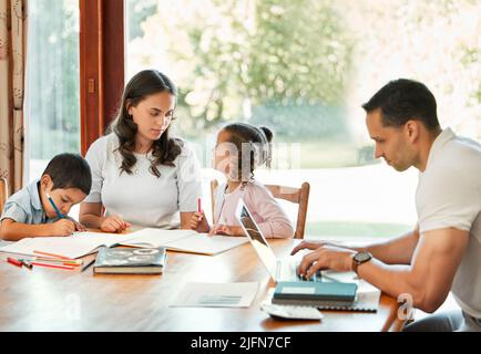 Jeune mère de race mixte aidant ses enfants à faire leurs devoirs pendant que leur père travaille sur un ordinateur portable à une table dans le salon. Dessin de petits frères et sœurs Banque D'Images