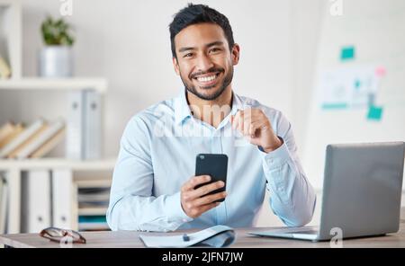 Portrait d'un jeune homme d'affaires de race mixte charmant utilisant les médias sociaux sur son téléphone tout en travaillant seul sur un ordinateur portable dans un bureau au travail. Un Banque D'Images
