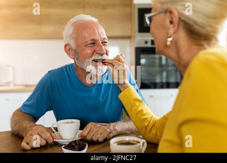 Femme nourrissant l'homme avec du chocolat à la table de salle à manger. Un couple senior boit du café à la maison Banque D'Images