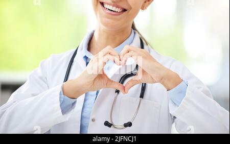 Femme inconnue médecin de race mixte assis dans son bureau et montrant un geste de forme de coeur avec sa main dans un hôpital. Une femme hispanique fait la main Banque D'Images