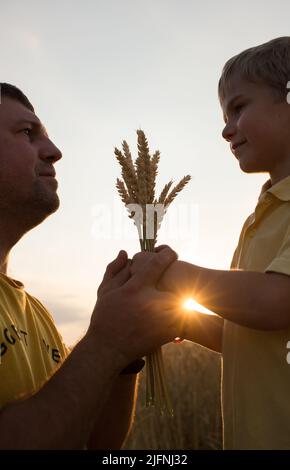 Silhouettes du visage et de l'enfant de l'agriculteur tenant un bouquet de spikettes entre les mains sur fond de soleil couchant. Récolte de céréales en Ukraine. Conc. Jour de la Terre Banque D'Images