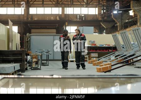 Deux jeunes travailleurs interculturels masculins de l'usine ayant une discussion des points de travail tout en se déplaçant le long d'un atelier spacieux Banque D'Images