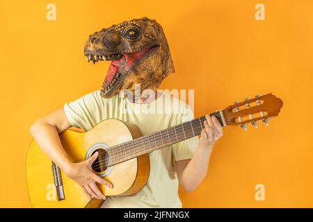 Portrait d'une femelle heureuse et méconnaissable dans un masque de dinosaure jouant de la guitare sur fond orange Banque D'Images