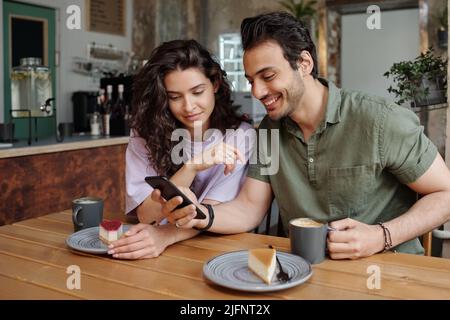 Jeune couple regardant à travers des photos drôles ou vidéo dans smartphone tenu par un gars tout en ayant cappuccino ou latte avec cheesecakes Banque D'Images