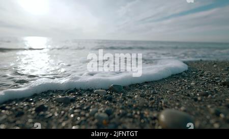 Gros plan sur les petites vagues. Action. Une petite vague roule sur la rive en pierre. Rive rocheuse lisse avec vague calme et vallonante le jour ensoleillé Banque D'Images