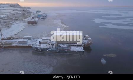 Vue aérienne de la zone industrielle d'une petite ville en hiver. Attache. Des arbres et des bâtiments enneigés et un quai près de la rivière. Banque D'Images