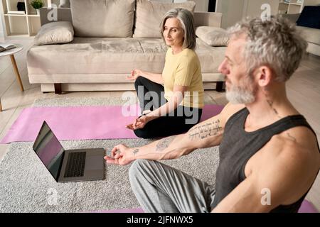 Un vieux couple d'âge mûr heureux faisant du yoga méditant ensemble à la maison avec un ordinateur portable. Banque D'Images
