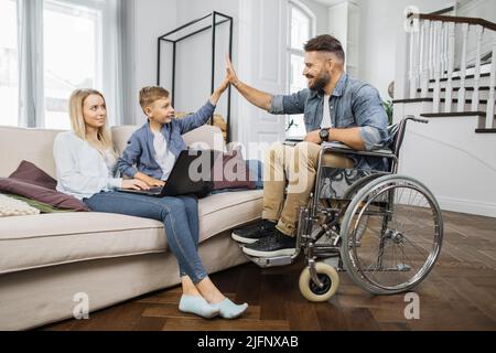 Bonne femme caucasienne assise avec son fils ensemble sur un canapé confortable et utilisant un ordinateur portable moderne. Enfant mignon faisant cinq grands gestes avec un père handicapé en fauteuil roulant. Banque D'Images