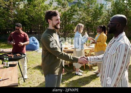 Deux jeunes hommes interculturels heureux se saluent en se faisant poignée de main contre deux femmes ayant une discussion par table de service Banque D'Images