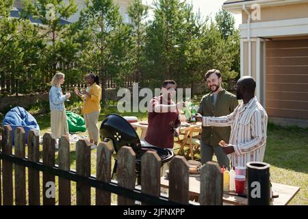 Trois jeunes hommes interculturels heureux se clinquant avec des bouteilles de bière pendant la fête en plein air le week-end tandis que l'un d'eux fait le barbecue sur le grill Banque D'Images