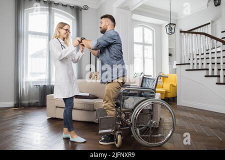 Jeune thérapeute positif aidant l'homme beau barbu à se lever du fauteuil roulant à la maison confortable. Un homme satisfait commence à sentir des muscles dans les jambes tout en essayant de marcher. Banque D'Images