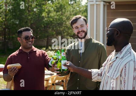 Groupe de jeunes hommes interculturels joyeux se clinquant avec des bouteilles de bière pendant la fête en plein air à la campagne et appréciant leur réunion Banque D'Images
