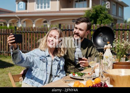 Un jeune couple ou des amis joyeux qui font du selfie sur un smartphone pendant un dîner en plein air ou une fête tout en étant assis près d'une table de service Banque D'Images