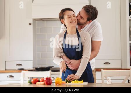 Baisers pour ma femme parfaite. Photo d'un homme qui embrasse sa femme pendant qu'elle prépare un repas dans la cuisine. Banque D'Images
