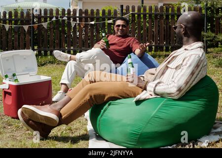Deux jeunes hommes interculturels avec des bouteilles de bière assis sur des fauteuils à sac et bavardant pendant la fête en plein air à la campagne le week-end Banque D'Images