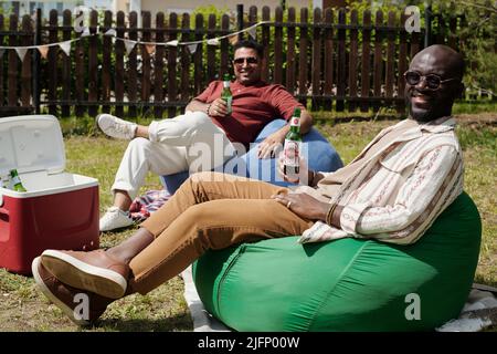 Deux jeunes hommes interculturels heureux avec des bouteilles de bière assis sur des chaises de sac et regardant la caméra pendant la fête en plein air le week-end Banque D'Images