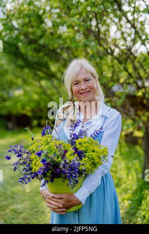 Bonne femme fleuriste senior portant un panier avec des fleurs à l'extérieur dans le jardin. Banque D'Images