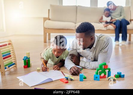 Jeune père afro-américain heureux aidant son fils à faire ses devoirs tout en étant assis sur le sol à la maison. Un petit garçon s'est concentré sur l'apprentissage et la réalisation d'une tâche Banque D'Images