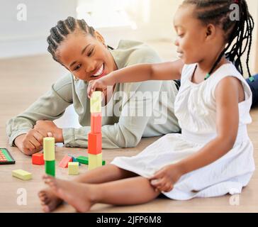 Fille afro-américaine jouant avec des blocs de construction tout en étant assise avec la mère. Petite tour de construction de fille avec des blocs de bois. Femme souriante couché Banque D'Images