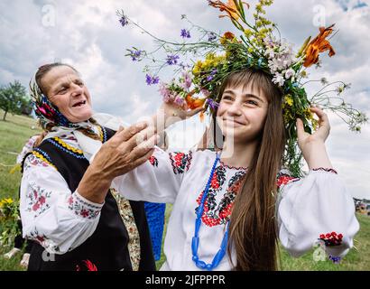 Une femme âgée vêtue de vêtements nationaux ukrainiens aide une fille dans une chemise brodée à mettre sur un headaddress de fleur pour la célébration. Cette fête symbolise la naissance du soleil d'été – Kupalo. L'histoire de l'Église chrétienne peut nous répondre comment la déité païenne Kupalo obtient le nom d'Ivan. Au IV ème siècle après J.-C., ce jour a été proclamé jour férié de la naissance de Jean-Baptiste - le précurseur de Jésus-Christ. À la suite de la Christianisation de la fête païenne, le nom 'Kupala' s'est associé au chrétien 'Ivan'. Sur le territoire de l'Ukraine pendant de nombreux siècles Kupala douanes cha Banque D'Images