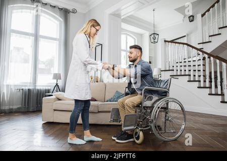 Jeune thérapeute positif aidant l'homme beau barbu à se lever du fauteuil roulant à la maison confortable. Un homme satisfait commence à sentir des muscles dans les jambes tout en essayant de marcher. Banque D'Images