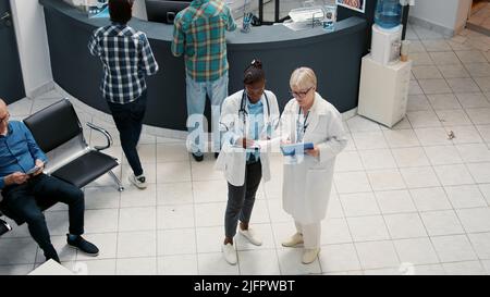 Équipe médicale diversifiée de médecins qui parlent et écrivent des rapports dans la zone d'accueil de l'hôpital, travaillant dans le système de soins de santé pour soutenir les patients malades. Rendez-vous dans le hall de la salle d'attente. Banque D'Images