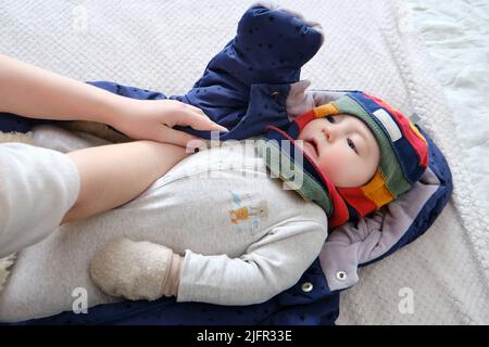 Une femme mère met une veste chaude pour un bébé garçon. Maman habille un enfant heureux dans des vêtements d'hiver sur le lit. Banque D'Images