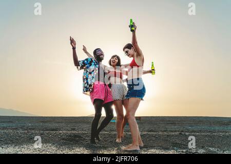 Joyeux groupe multiracial de jeunes dansant fou sur la plage au coucher du soleil boire des bières en bouteille - personnes multiethniques, amusement, amitié alcool et Banque D'Images
