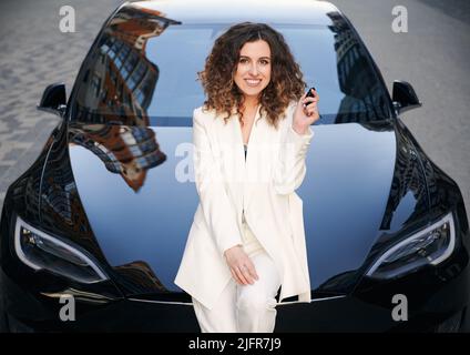 Portrait d'une femme d'affaires élégante tenant la clé de la voiture, se penchant contre le pare-chocs avant de l'automobile. Belle dame aux cheveux bouclés en costume blanc souriant sur le fond d'une voiture de luxe chère. Banque D'Images