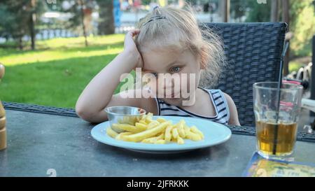 Une petite fille mange des frites. Gros plan de blonde fille prend des chips de pomme de terre avec ses mains et les essaie assis dans la rue café sur le parc. Banque D'Images