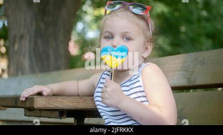 Petite fille blonde tient le pain d'épice dans les couleurs nationales ukrainiennes dans sa main, il dit «l'Ukraine est moi». Portrait en gros plan d'une fille assise sur le parc Banque D'Images