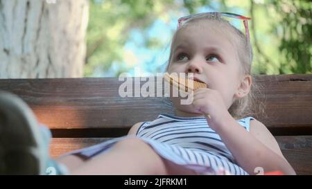 La petite fille mange du pain d'épice. Portrait en gros plan d'une fille blonde assise sur le banc du parc et mangeant des biscuits. Banque D'Images